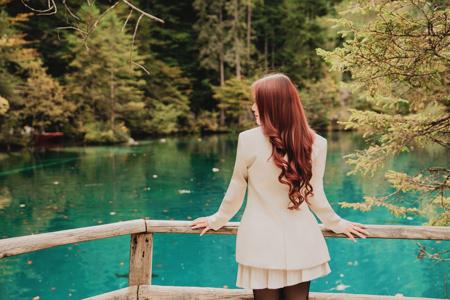 Young woman with long red hair in white outfit overlooking stunning turquoise mountain lake.