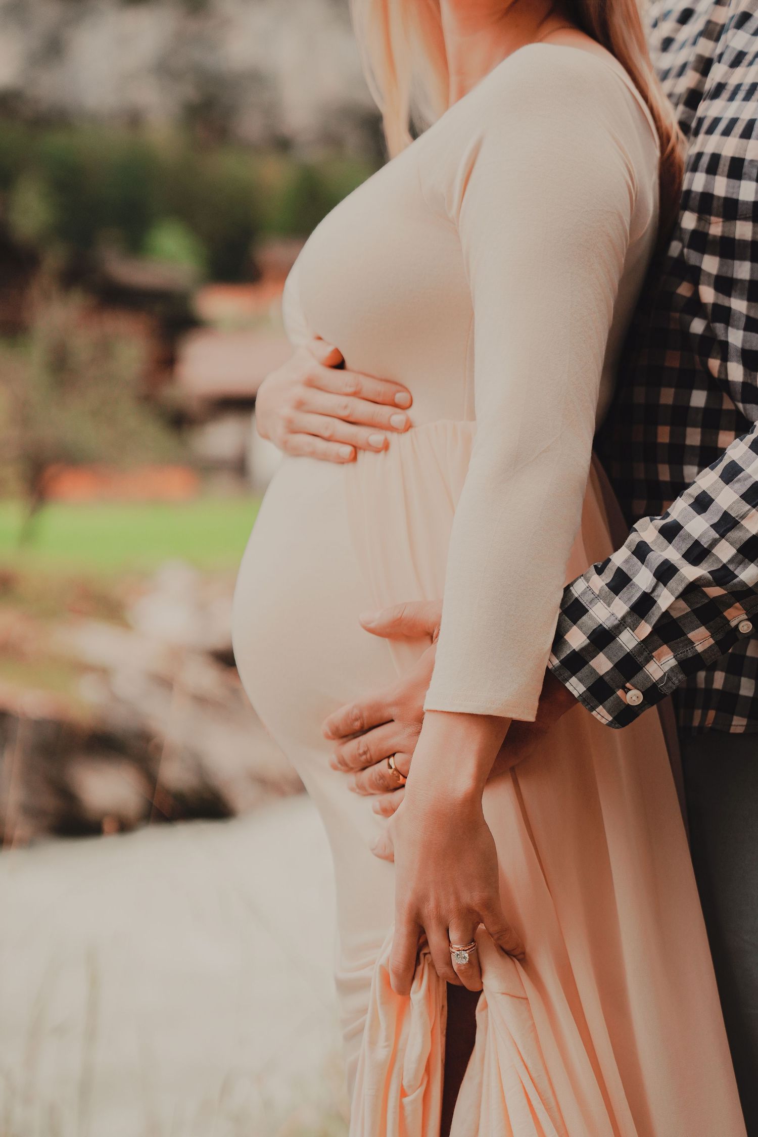 Close-up of pregnant belly with couple tenderly holding bump in soft blush maternity dress outdoors.