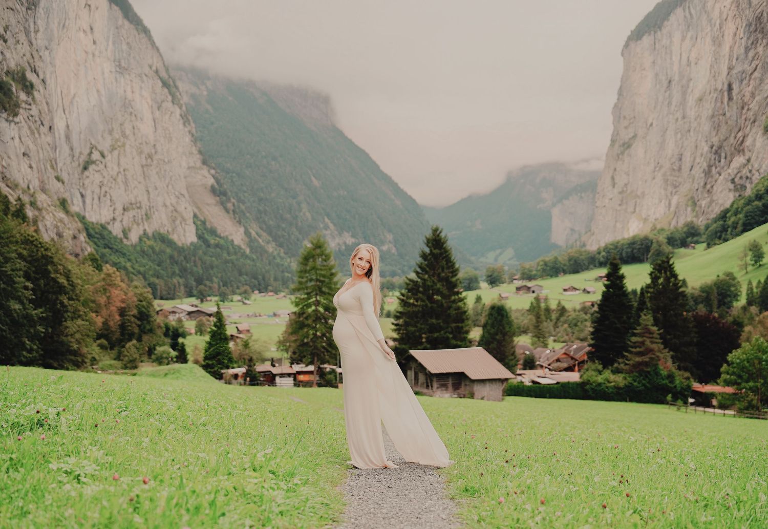 Pregnant blonde woman in cream maternity gown standing on a gravel path in a wide Swiss alpine valley.
