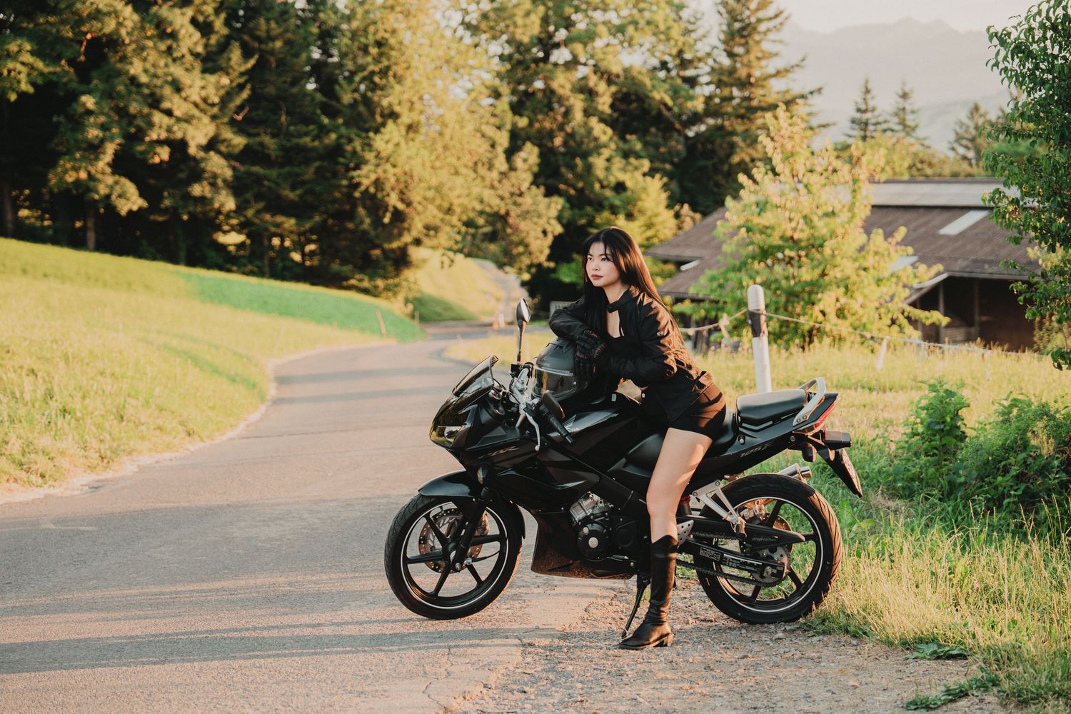 Young woman in black outfit posing beside a black sport motorcycle on a scenic rural road at golden hour.