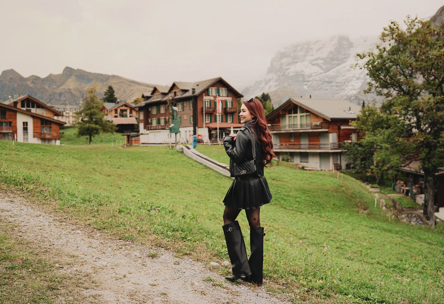 Woman in black outfit stands on a grassy hillside path surrounded by traditional Swiss Alpine chalets.