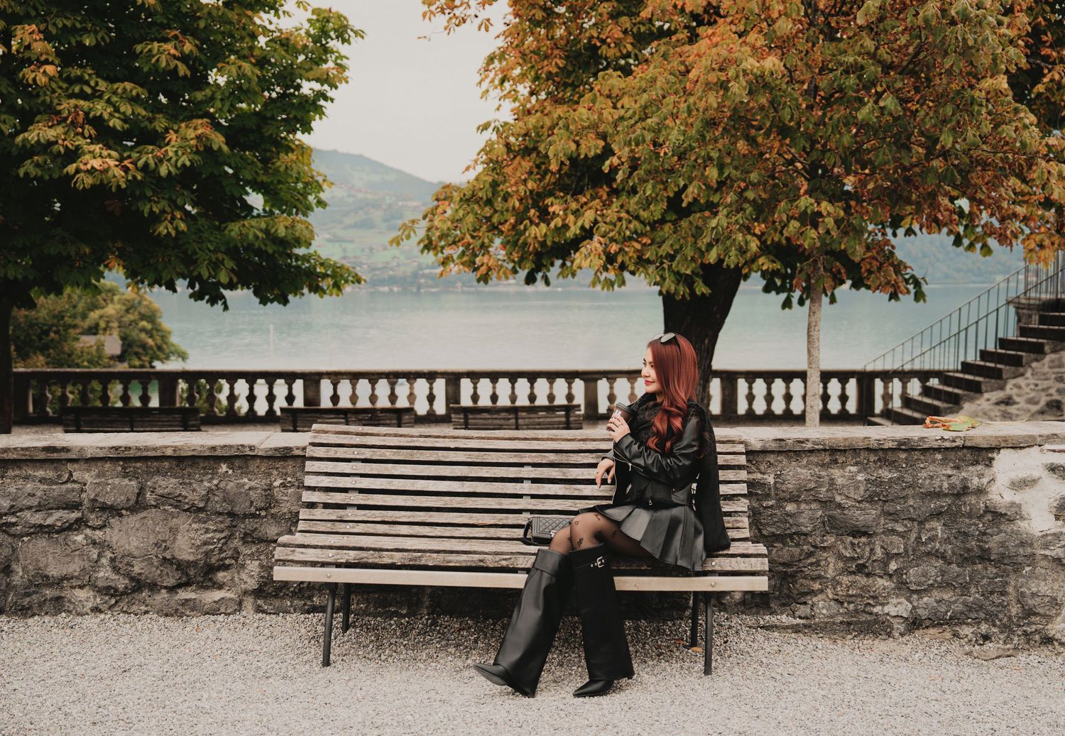 Red-haired woman seated on wooden bench in autumn park with lake and colorful foliage behind her.