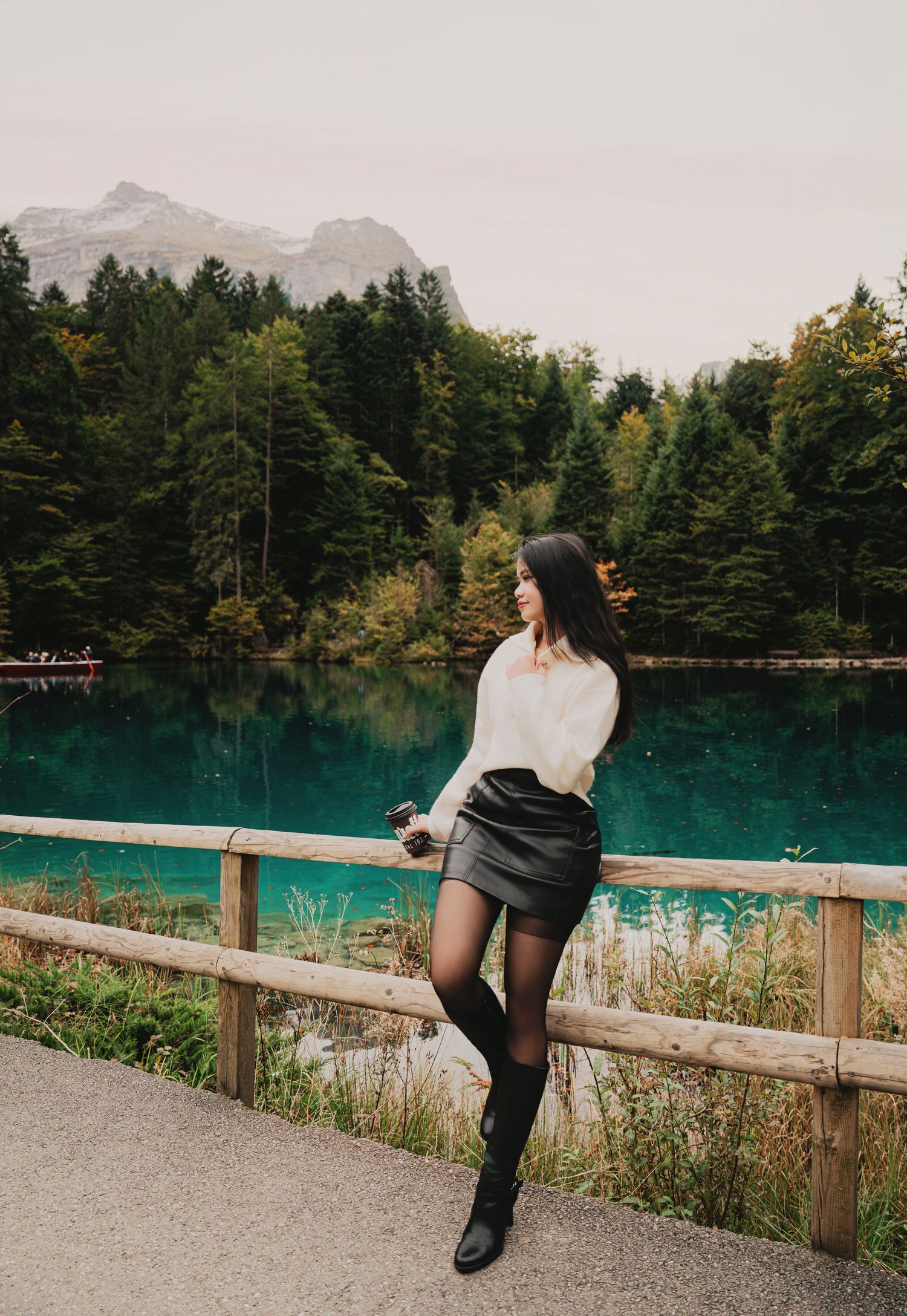 Woman in white sweater and black mini skirt with tall boots posing by turquoise forest lake.
