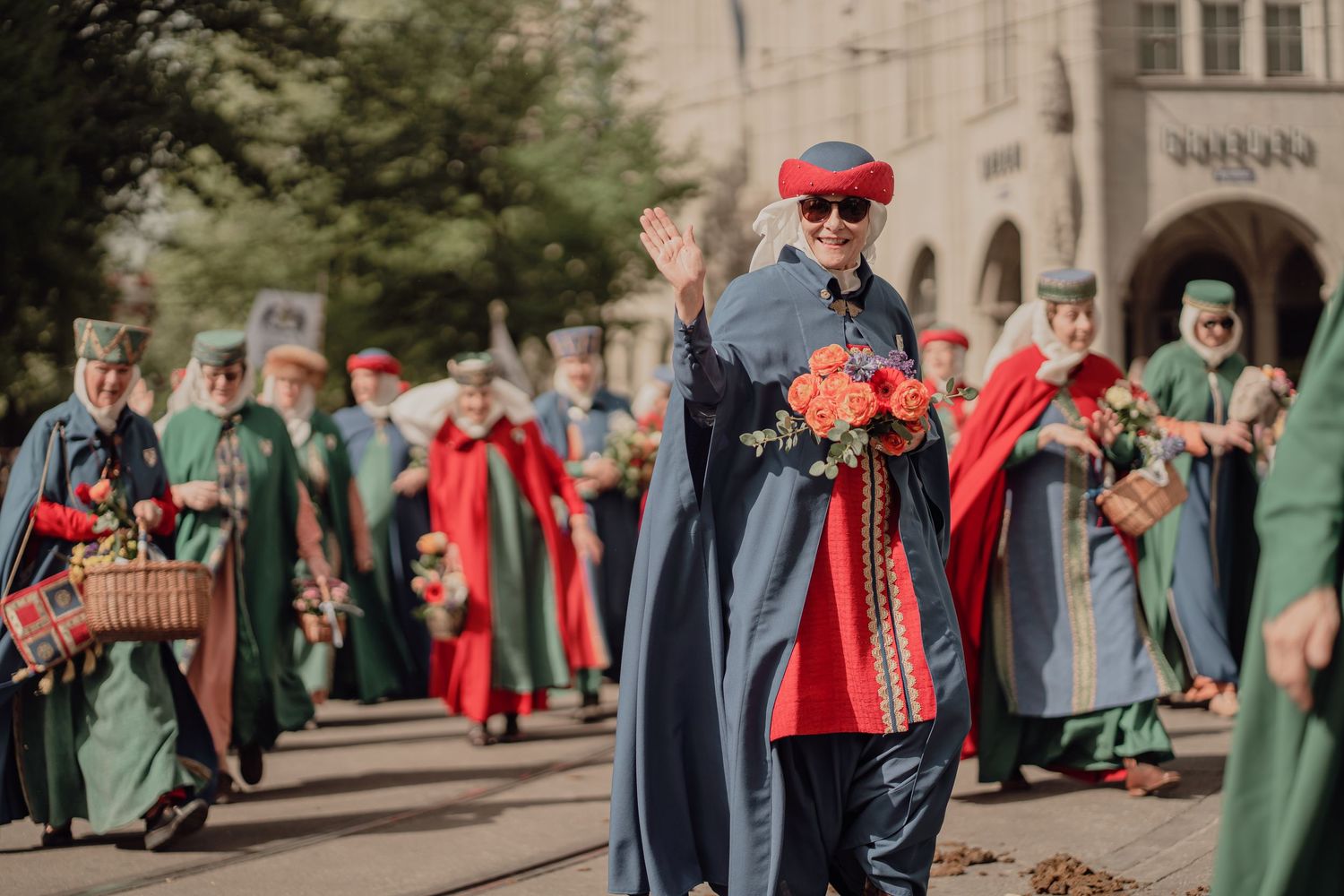 Smiling person in blue and red medieval costume waves while holding roses in a festive parade.