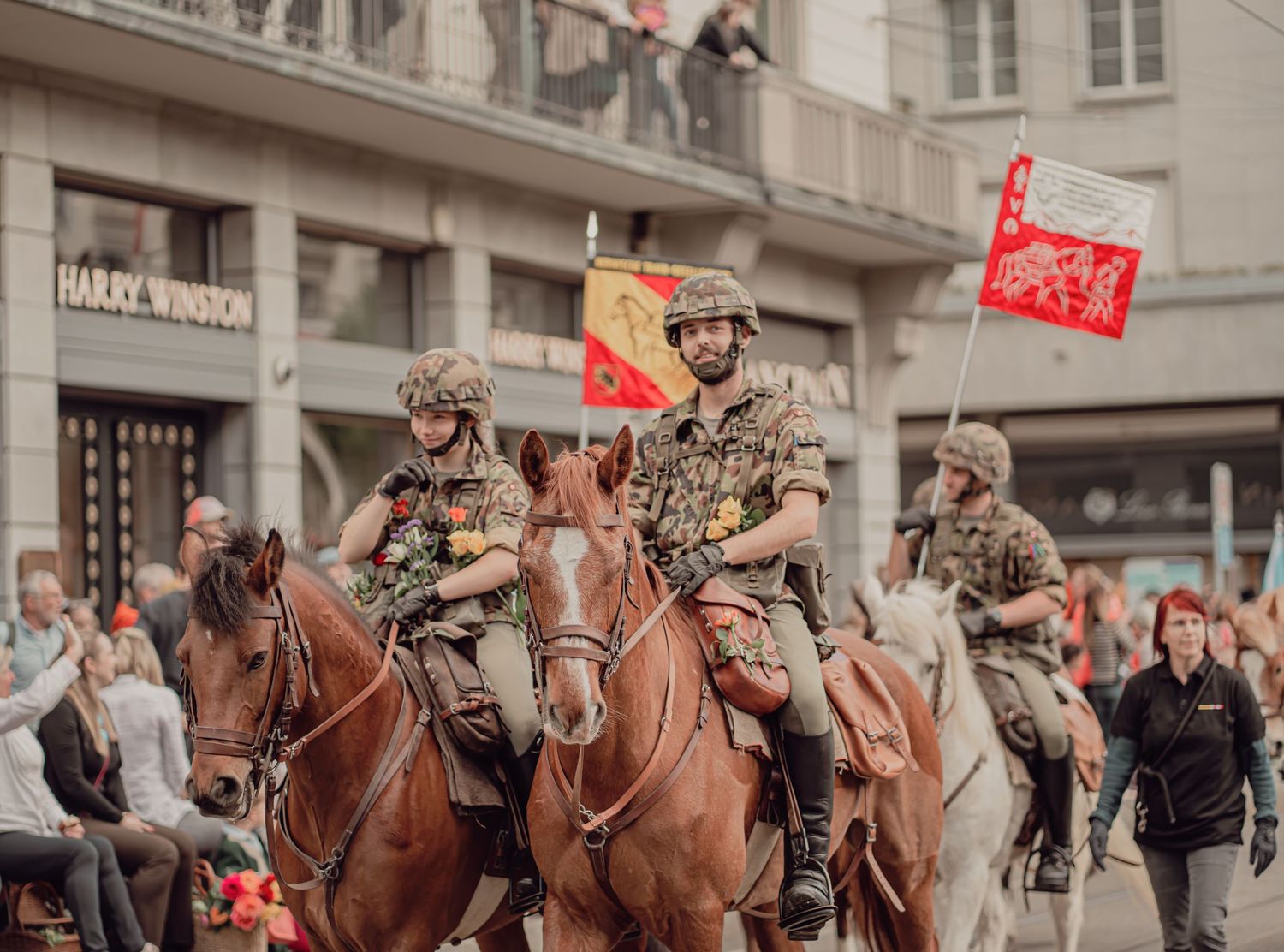 Uniformed riders on brown horses wave flags during a lively city parade event.