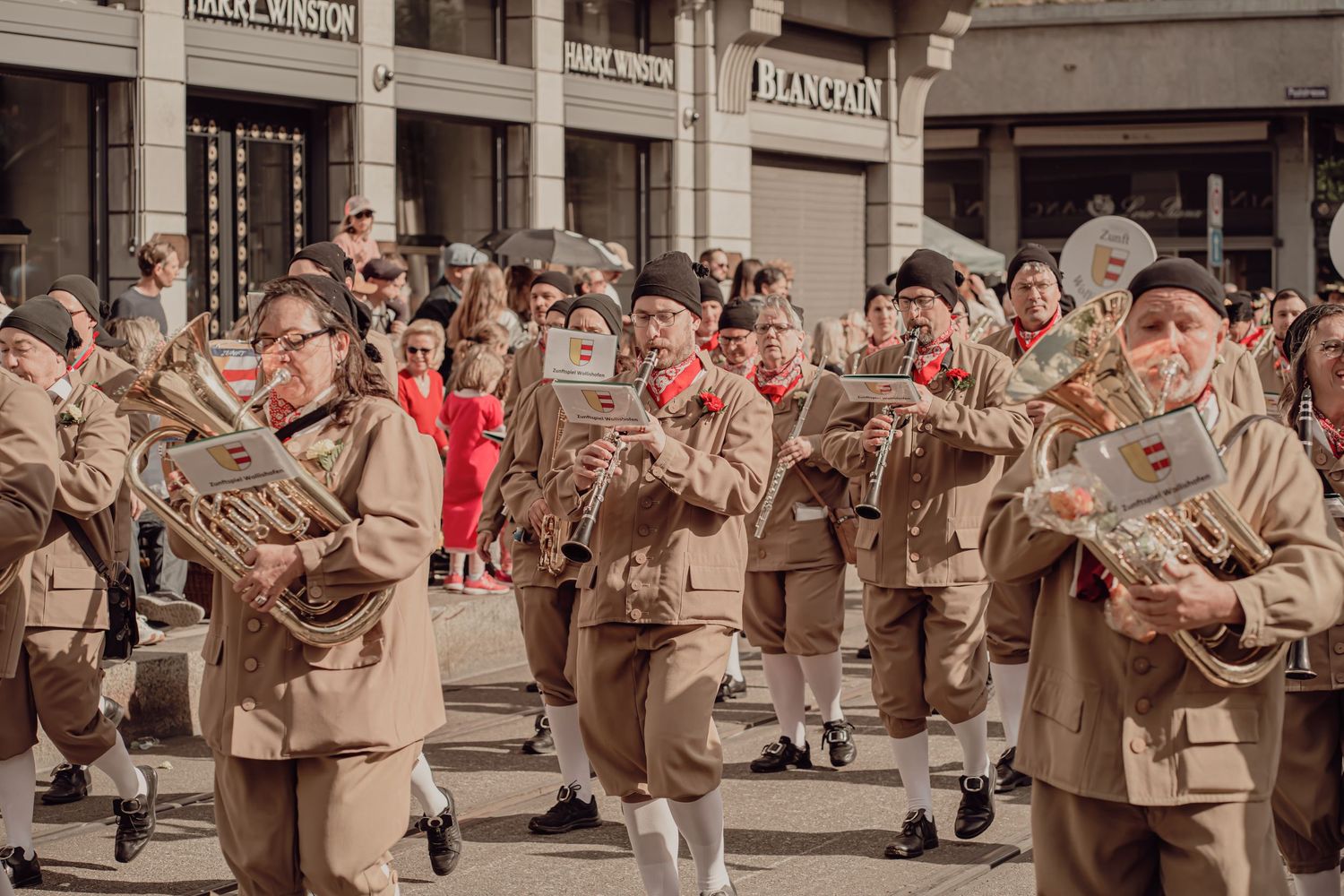 Marching band in matching tan uniforms plays brass instruments during a festive street parade.