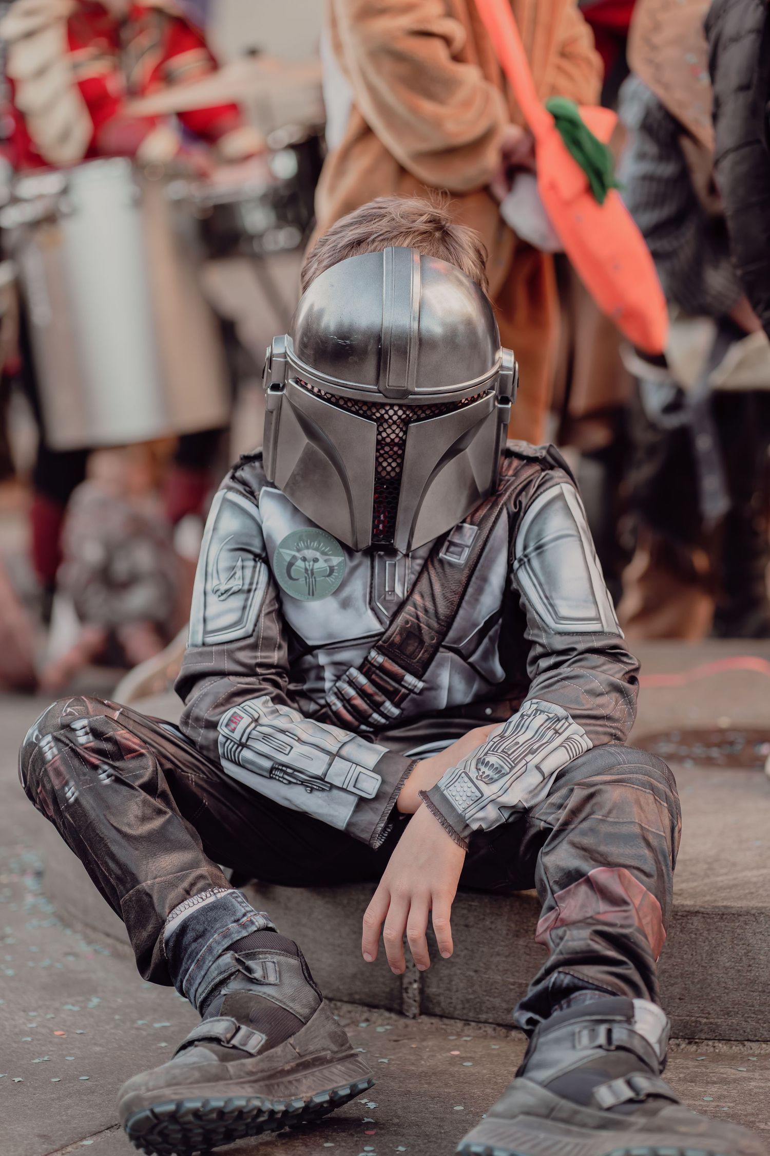 A child dressed in a Mandalorian Star Wars costume sits on a curb at a carnival.