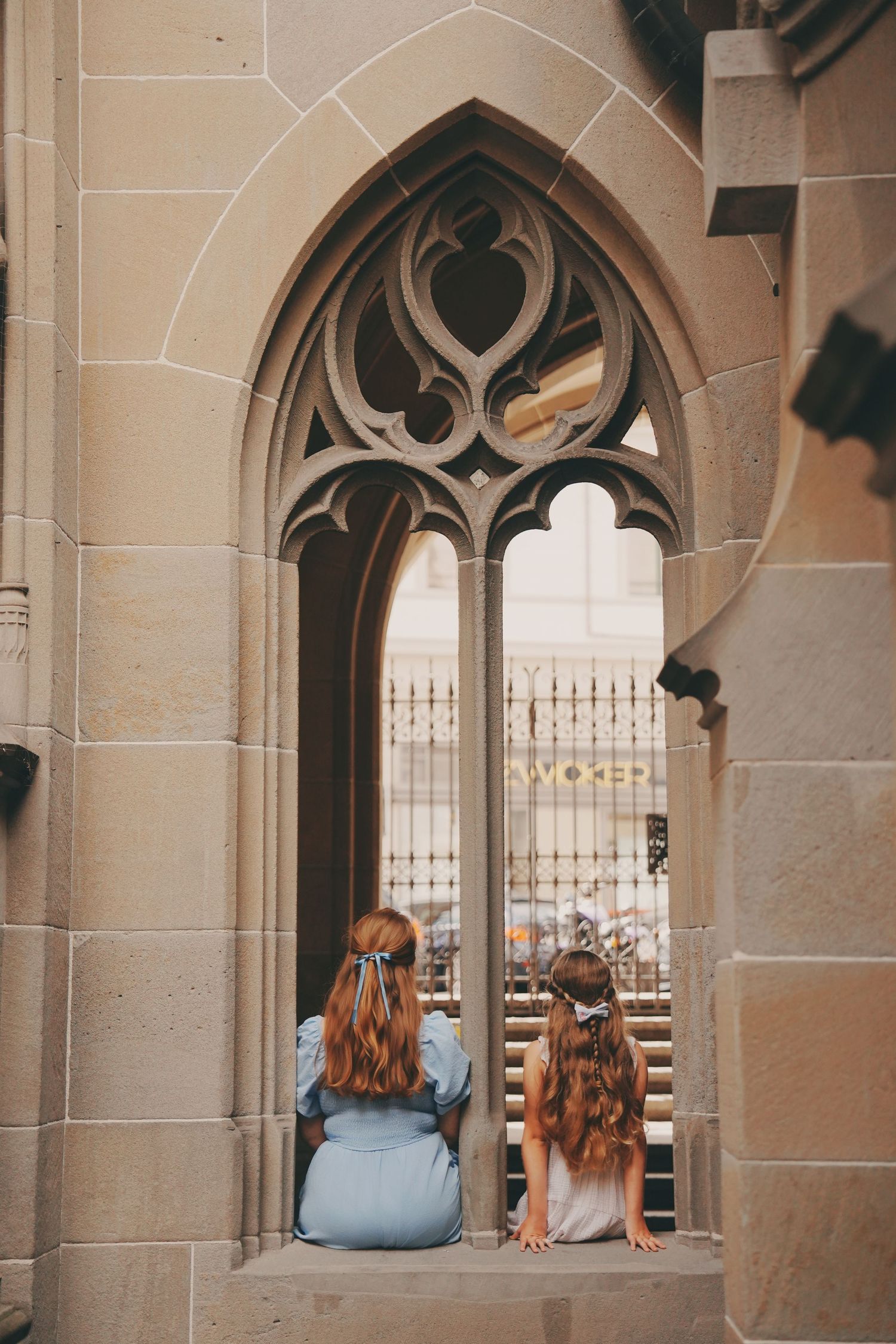Two girls with long hair sitting in ornate Gothic arched window of historic stone church.