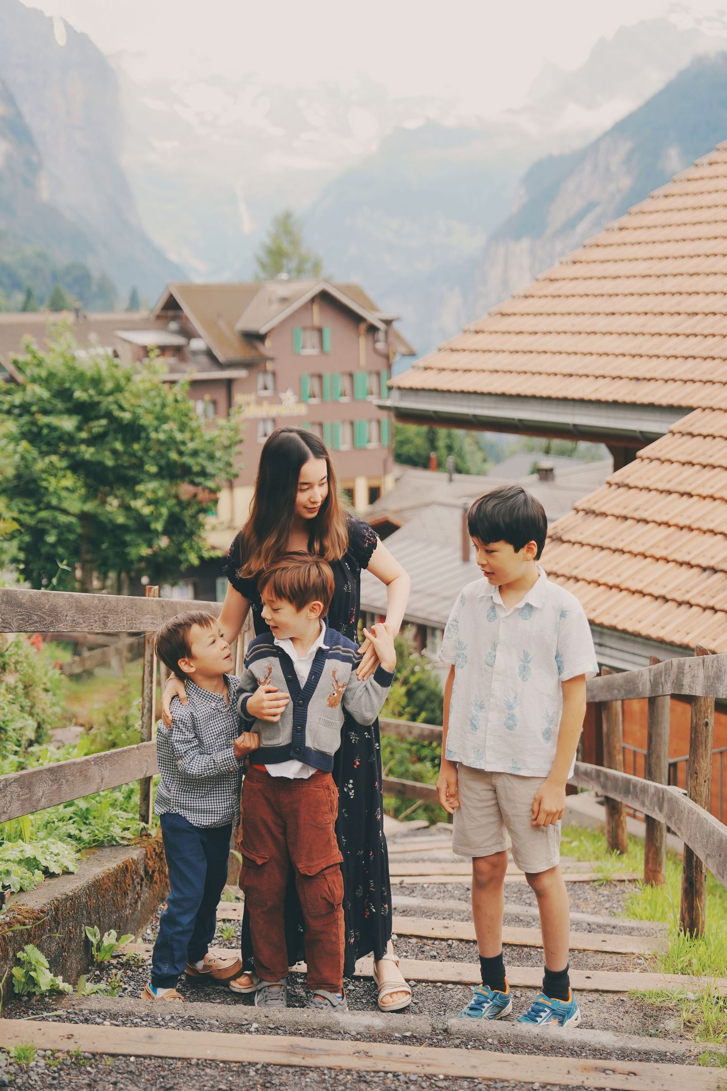 Asian mother embraces three sons on wooden stairs with majestic snow-capped Swiss Alps in background.