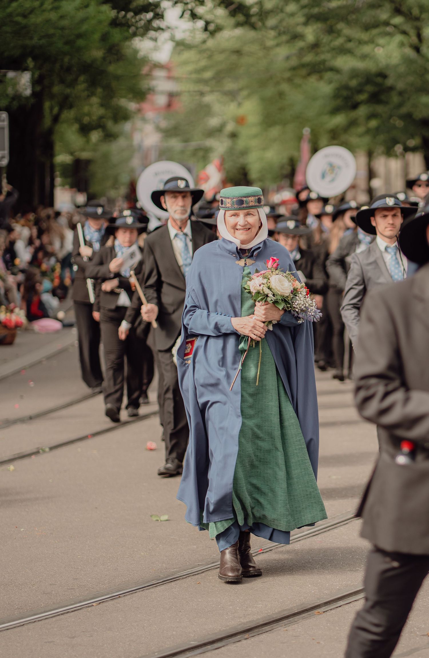 A smiling woman in a blue and green historical costume holds flowers while marching in a city parade.