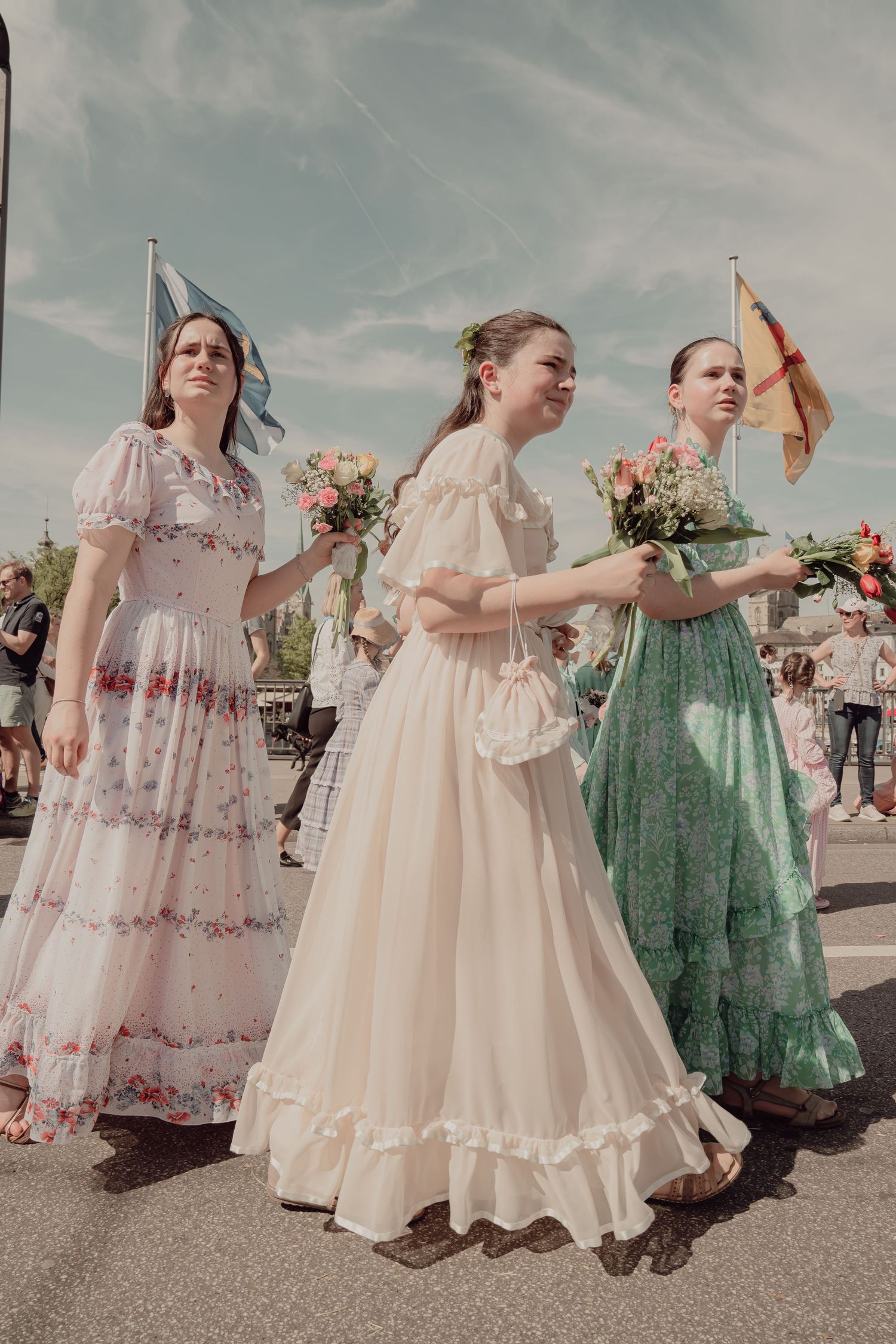 Three women in elegant long floral and pastel dresses carrying bouquets in a traditional Swiss parade.