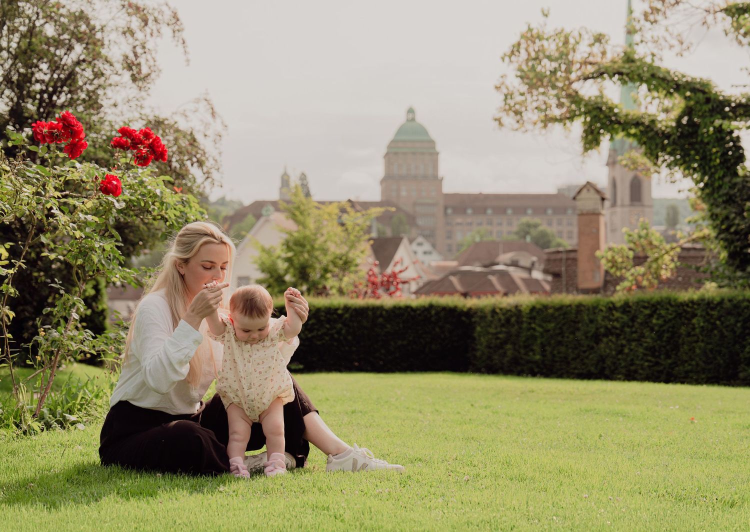 Mother and baby girl sitting on green lawn with red roses and historic building skyline.