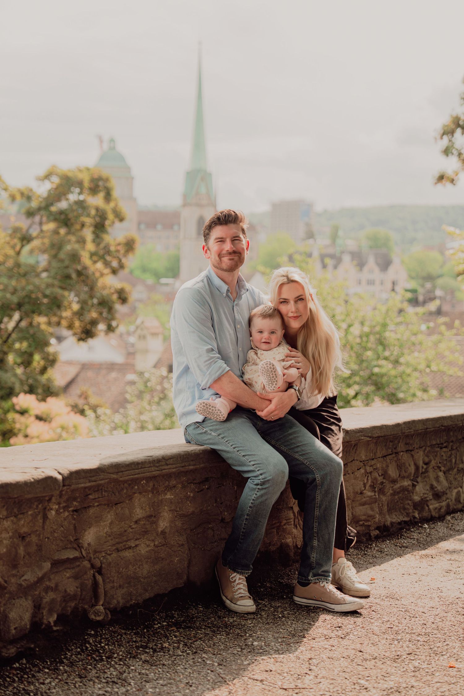 Young family with baby sitting on stone wall, historic European city with church spire visible behind.