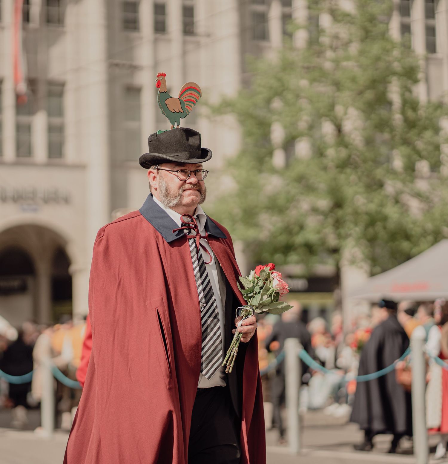 Elderly man in red cape and bowler hat with rooster decoration holds roses at an outdoor event.