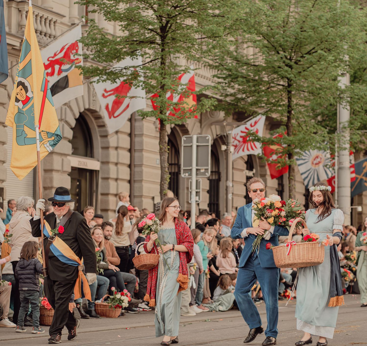 Festival participants in period costumes carrying flowers and flags past a crowd of spectators.