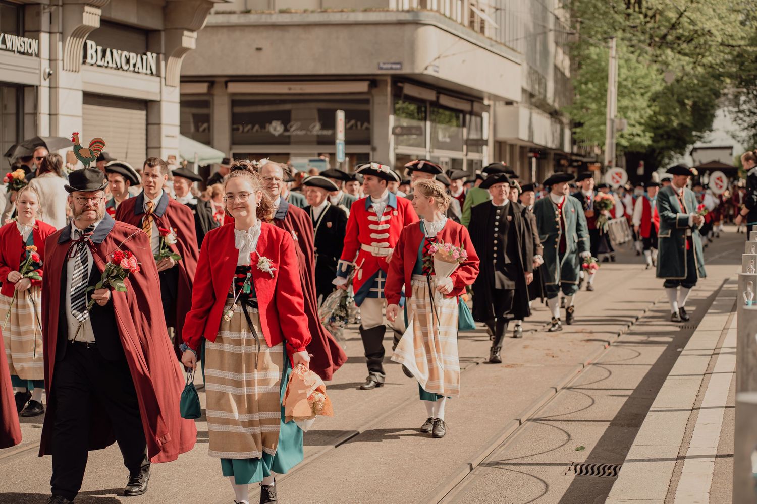 Crowd of participants in red historical uniforms and traditional dress marching through a city street parade.