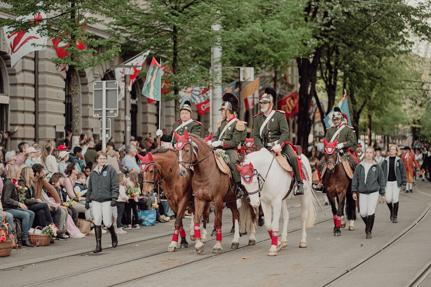 Riders in historical uniforms on horseback parade before a large crowd lining a city boulevard.
