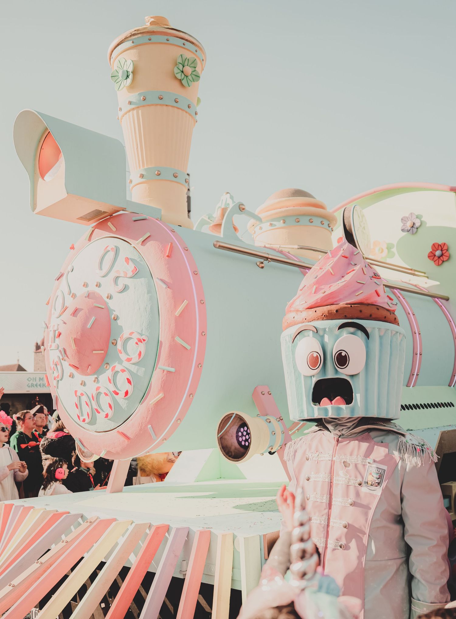 Pastel candy-decorated train float with a cupcake mascot costume character at a colorful parade.