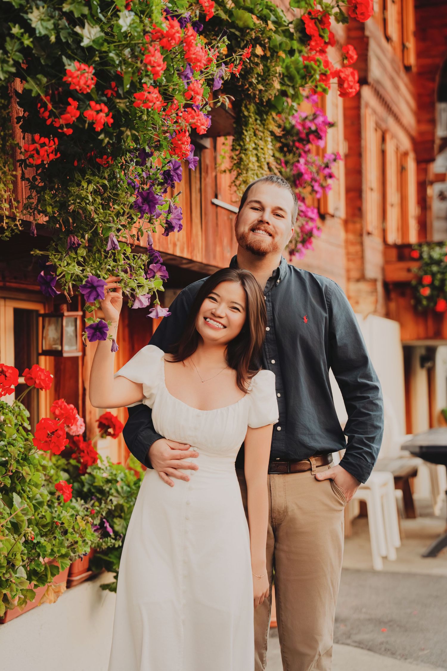 Smiling couple posing beside a Swiss building adorned with colorful red and purple flowers.
