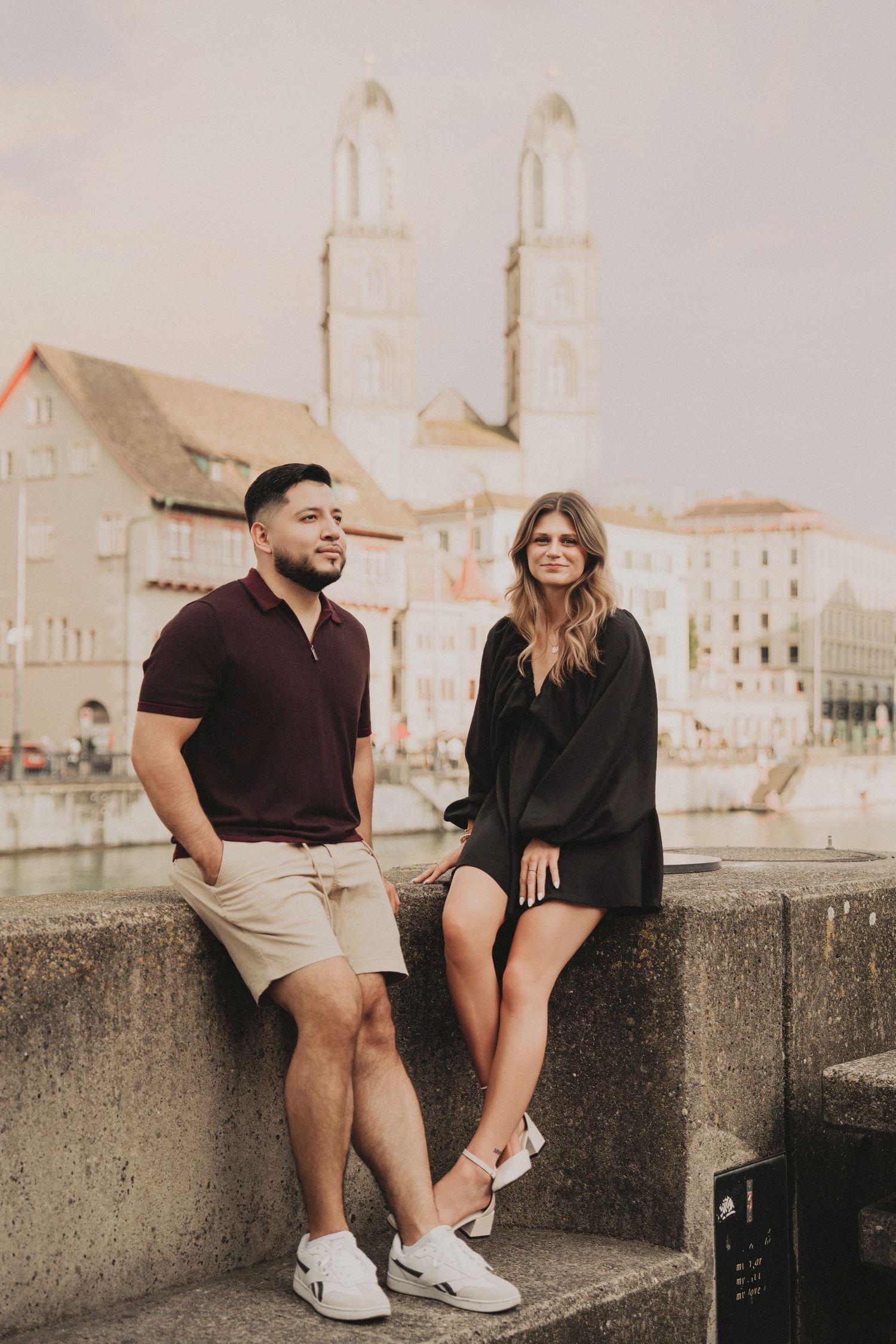 Stylish couple seated on a stone bridge railing with Zurich's iconic Grossmunster cathedral in the background.