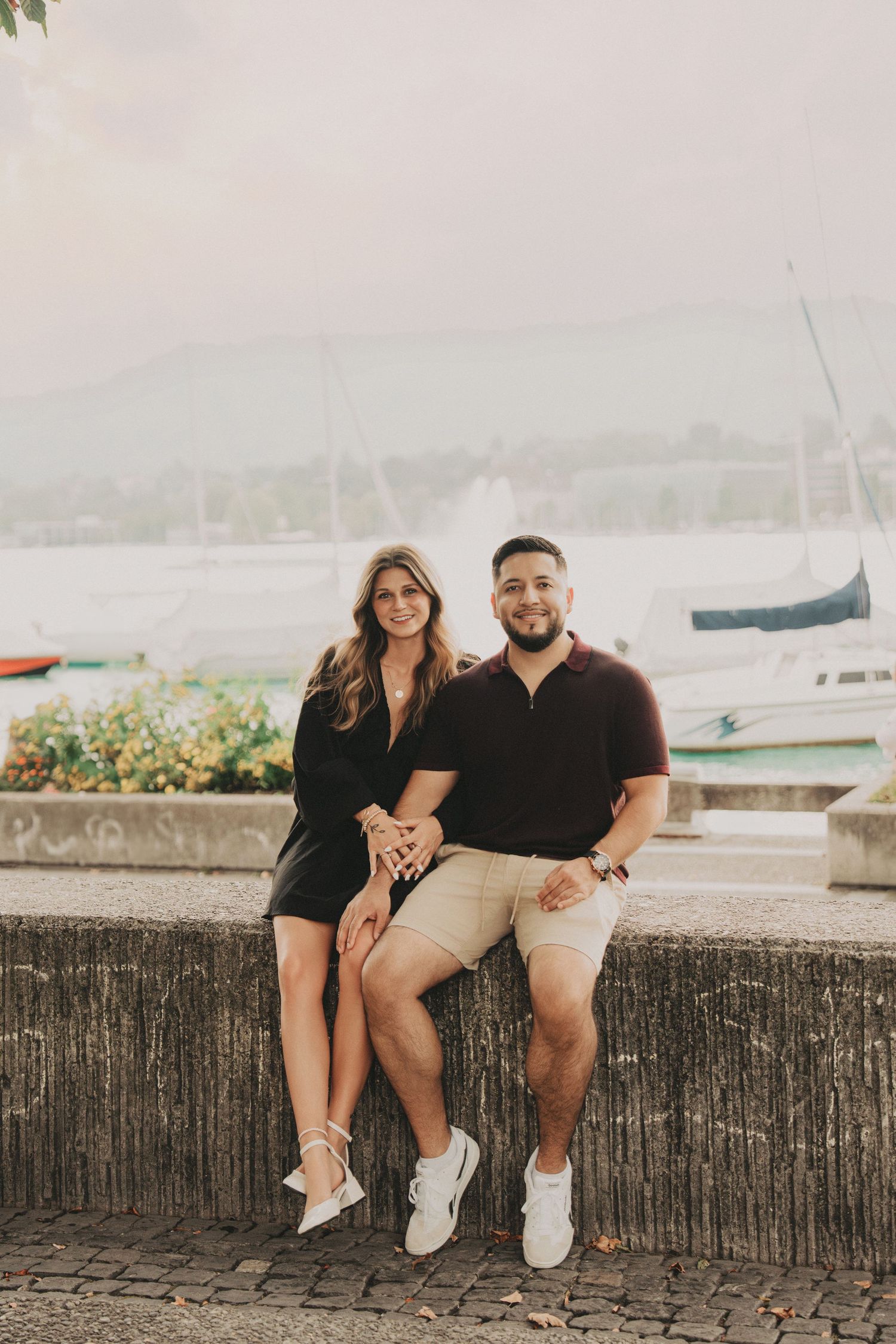 Smiling couple seated on a stone wall with sailboats and mountains in the background in Zurich.