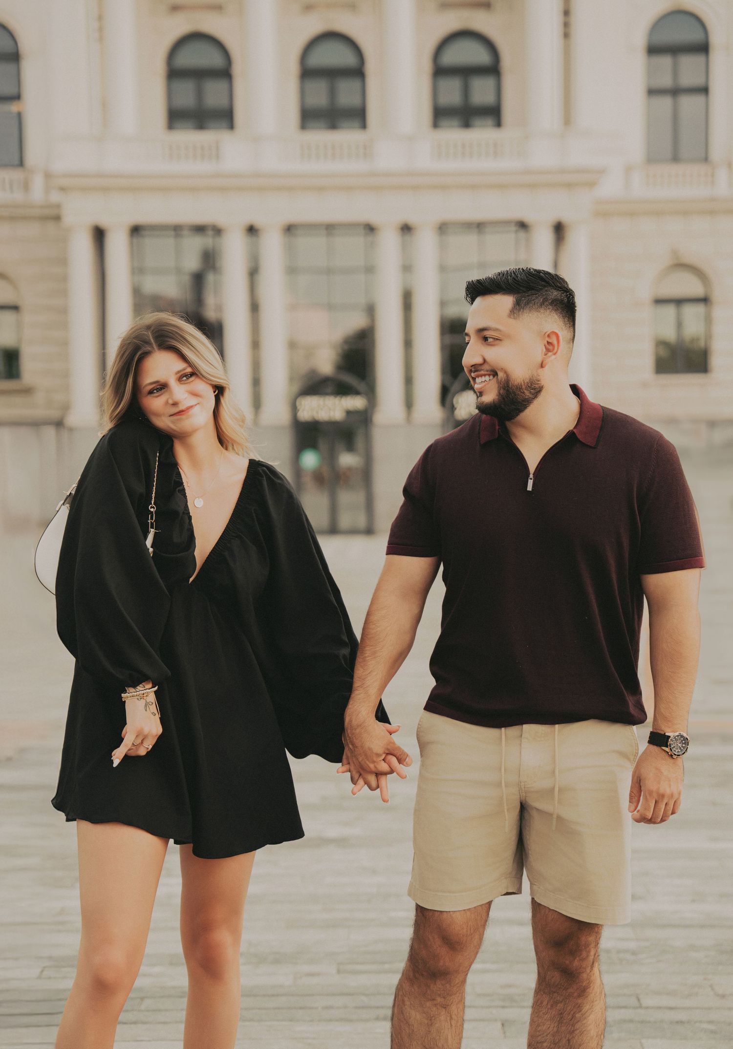 Smiling couple in black and tan outfits walking hand in hand near a historic European building.