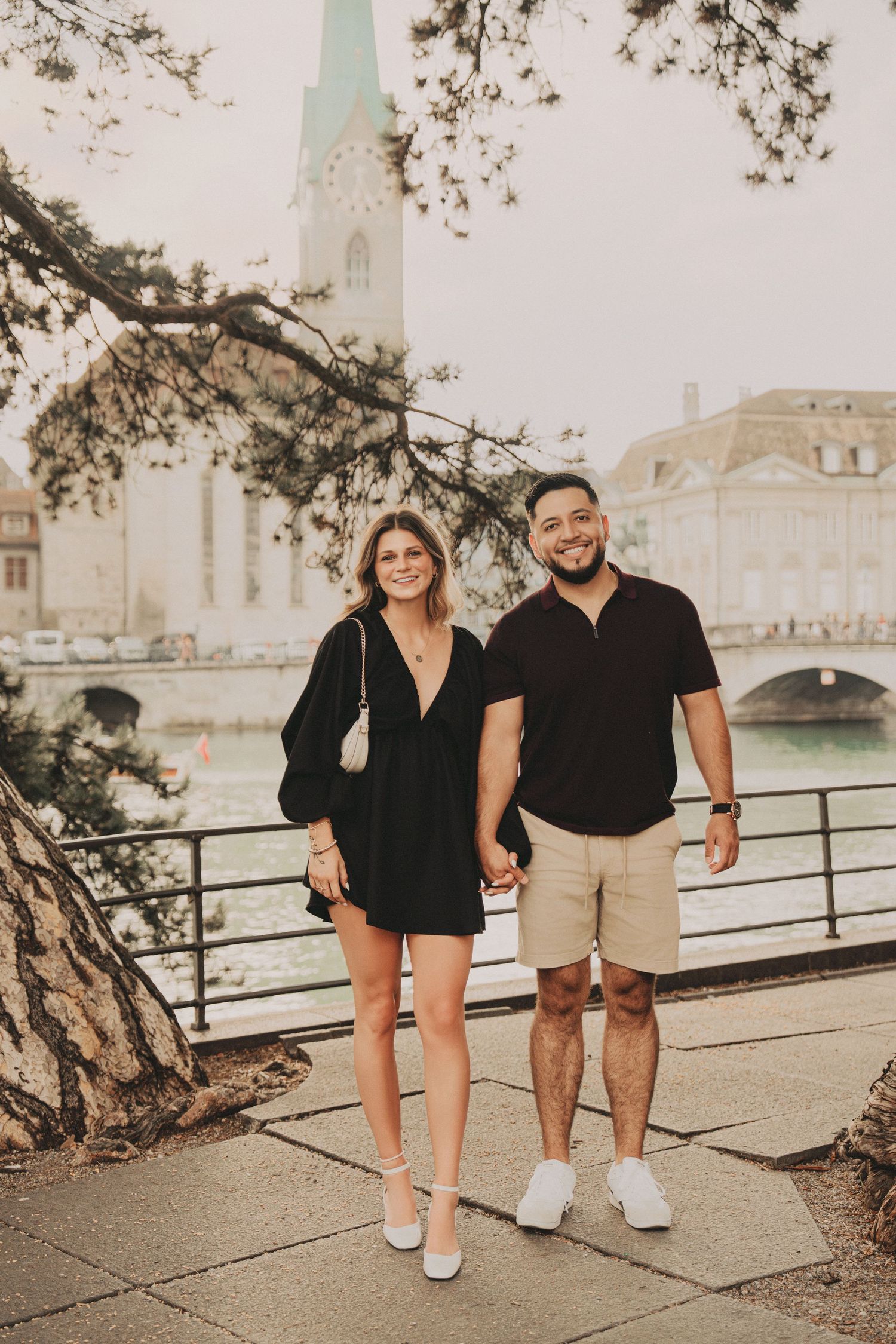 Happy couple posing hand in hand along Zurich riverside with church spire and historic buildings.