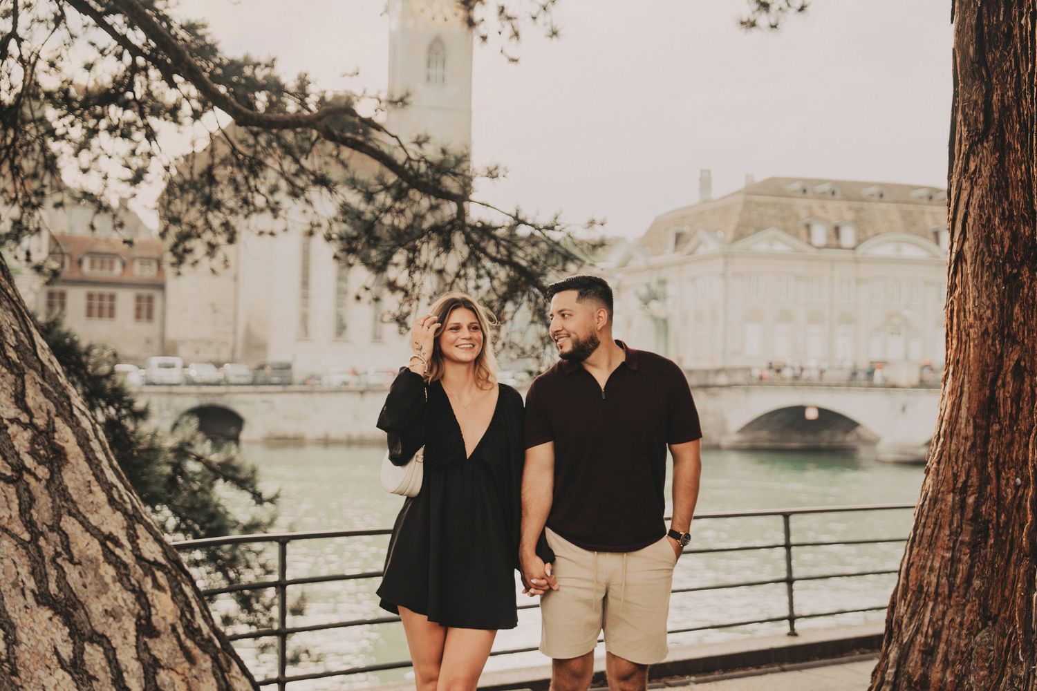 Couple walking hand in hand along Zurich riverside with historic buildings and bridge behind them.