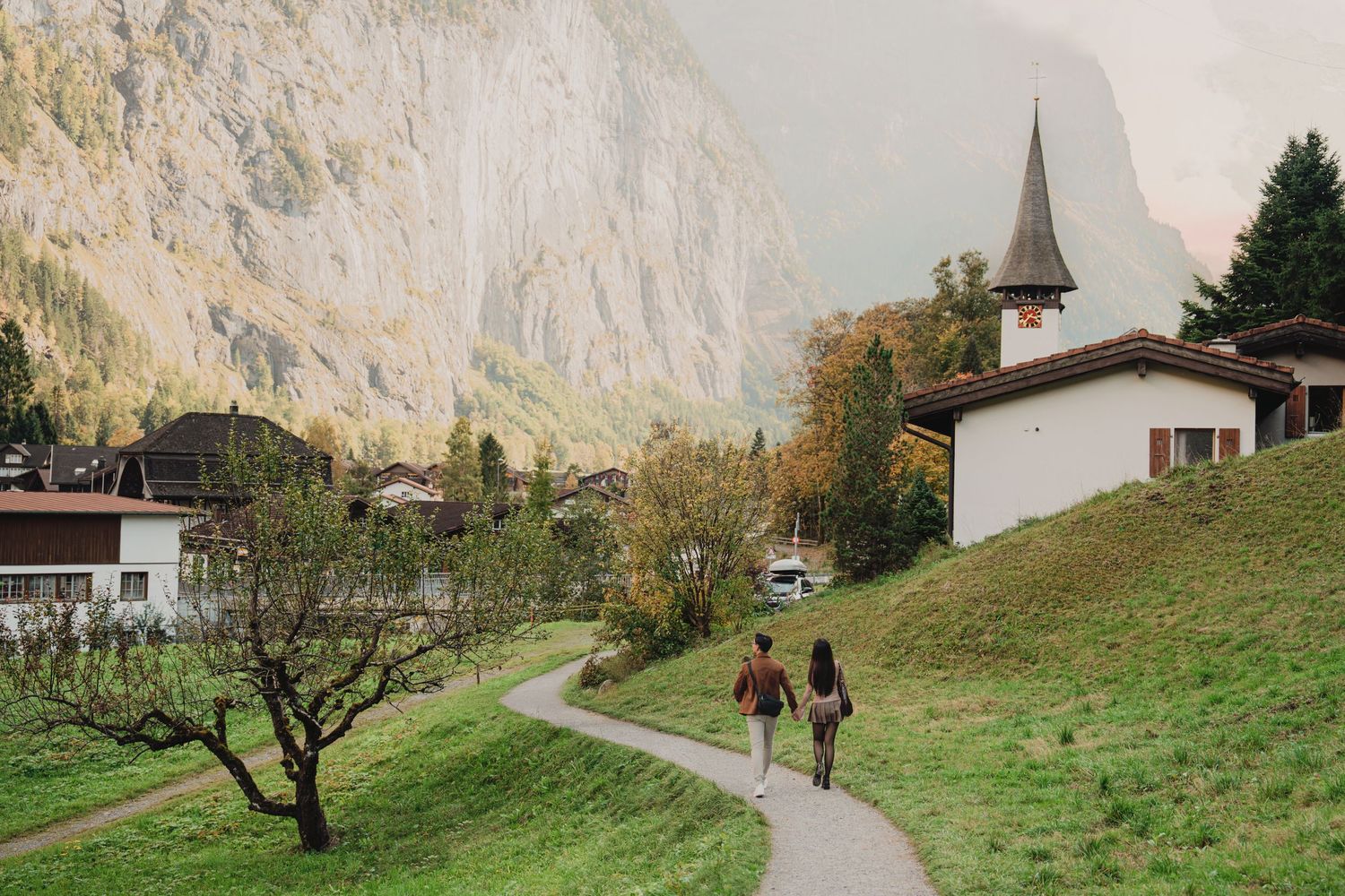Two people strolling along a winding path through a charming Swiss alpine village near a church.