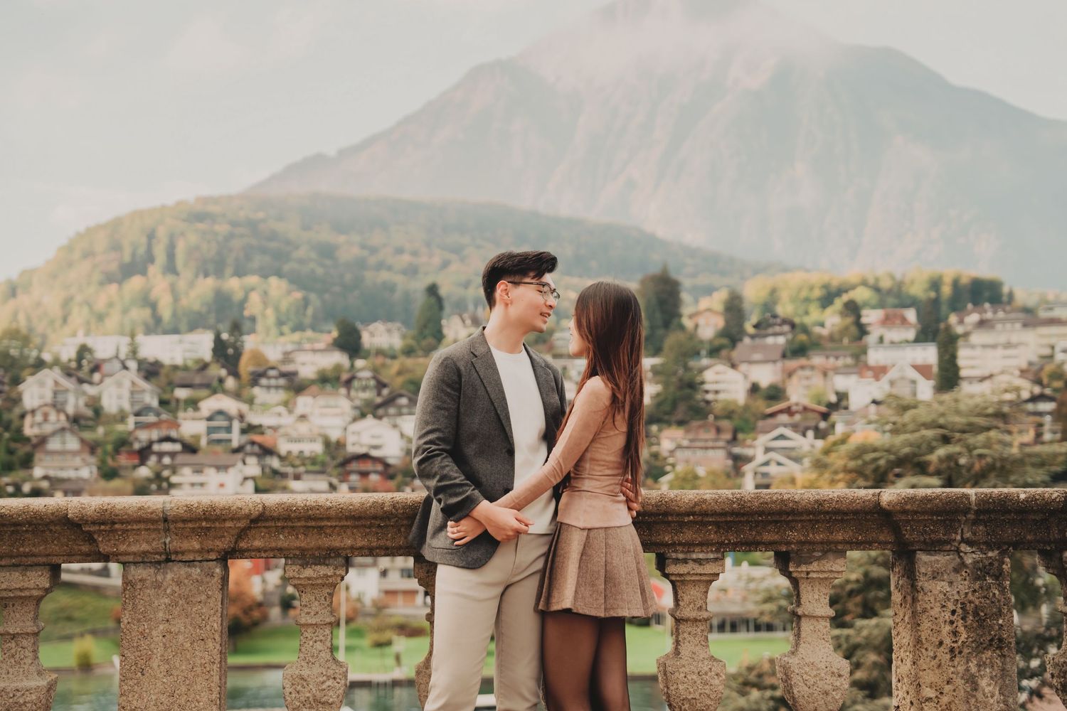 Couple gazing lovingly at each other on elegant terrace with European mountain village behind them.