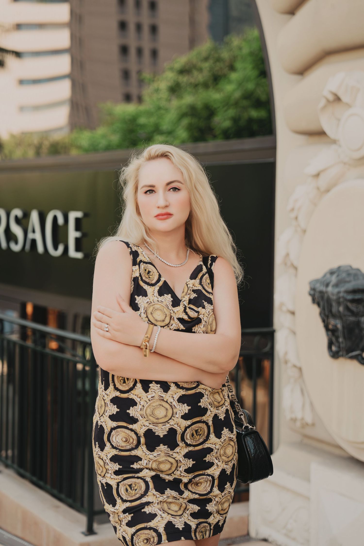 Blonde woman in black and gold Versace dress posing with arms crossed outside Versace store.