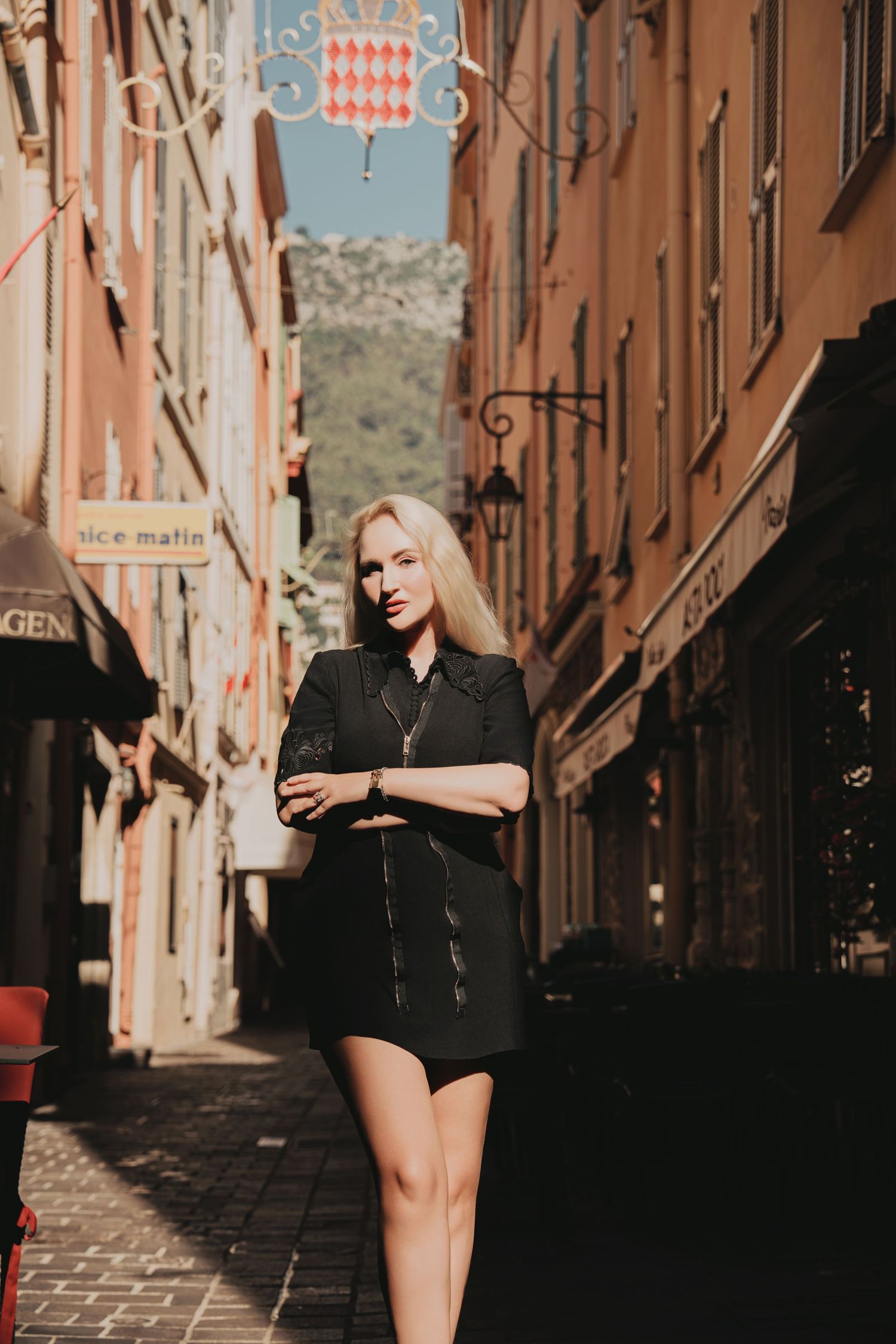Blonde woman in black zip dress with arms crossed in Monaco old town alleyway.