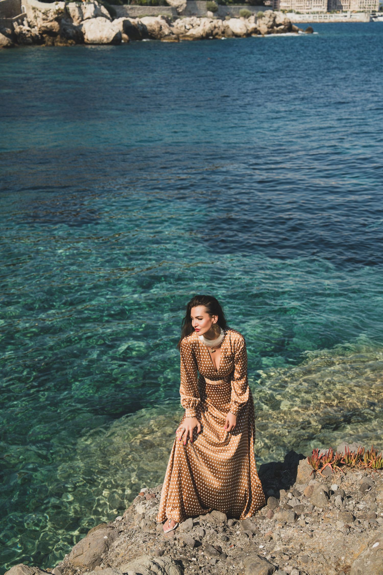 Woman in gold polka dot maxi dress poses on coastal rocks with turquoise Mediterranean sea backdrop.