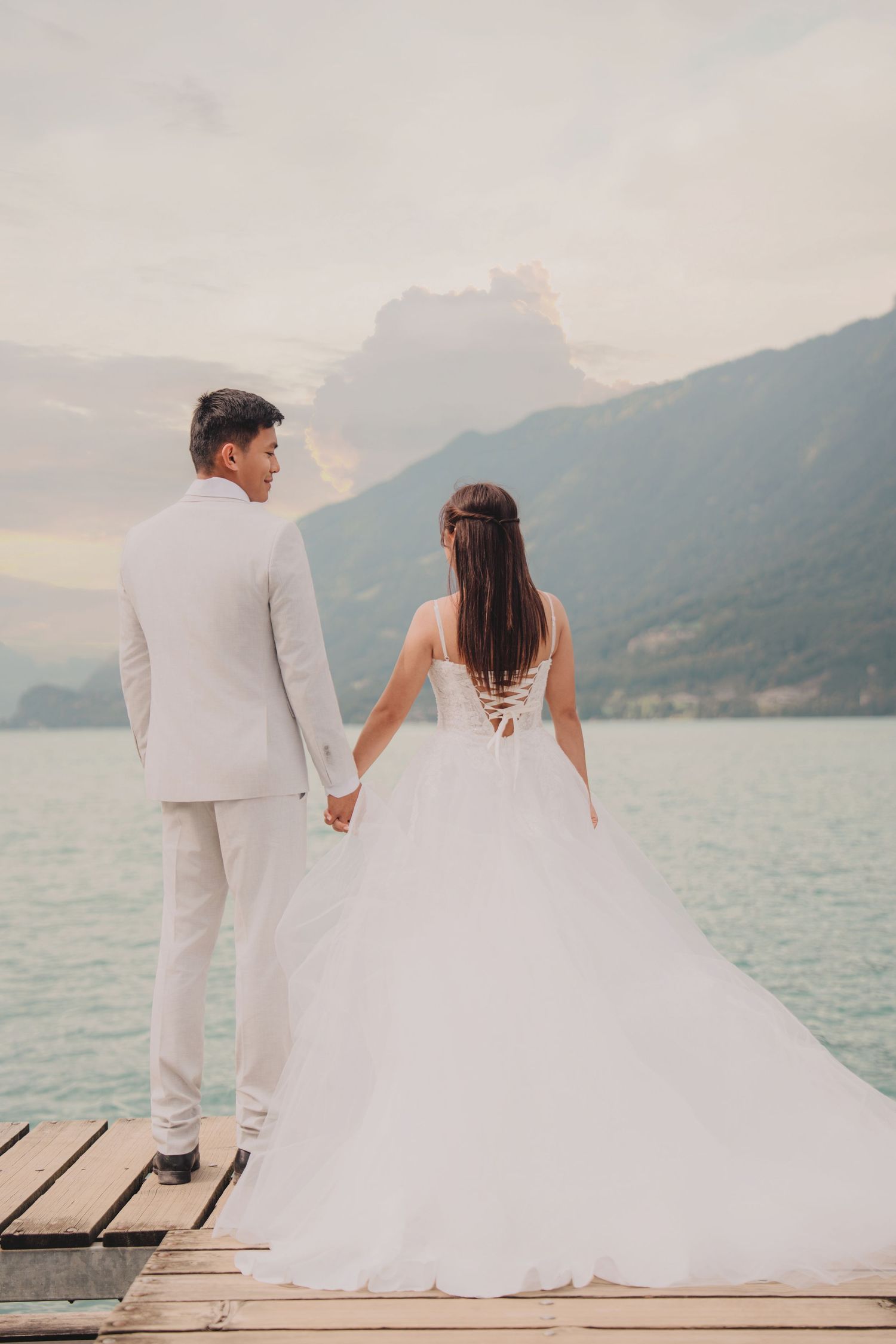 Bride and groom hold hands on wooden pier, bride in lace-back gown, overlooking alpine lake and mountains.