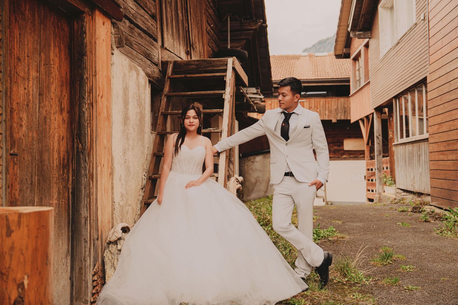 Bride and groom walk playfully hand in hand past rustic wooden barn buildings on wedding day.