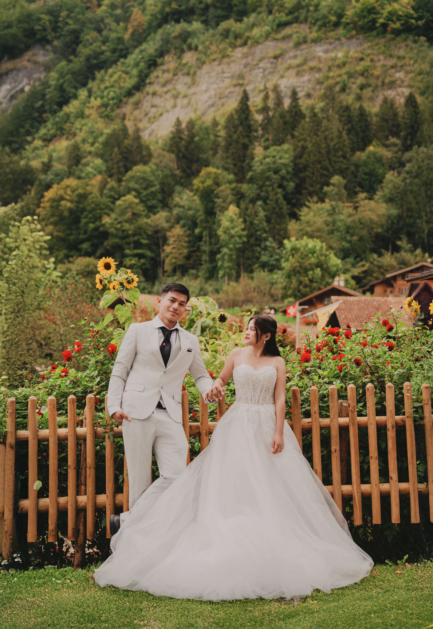 Wedding couple holding hands in flower garden with sunflowers, wooden fence, and forested alpine hills.