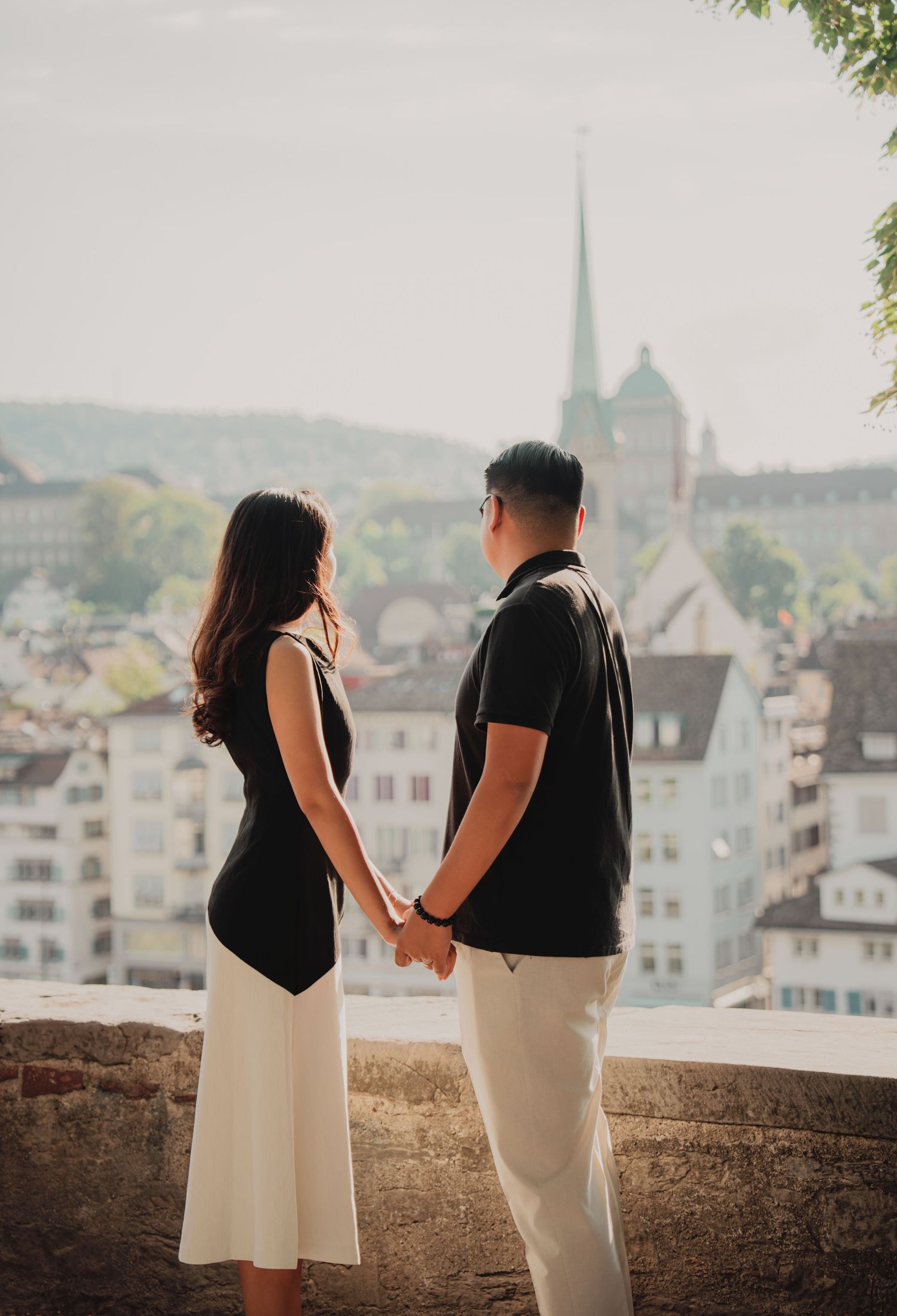 Couple holding hands while gazing at the Zurich skyline featuring a church spire from a hilltop viewpoint.