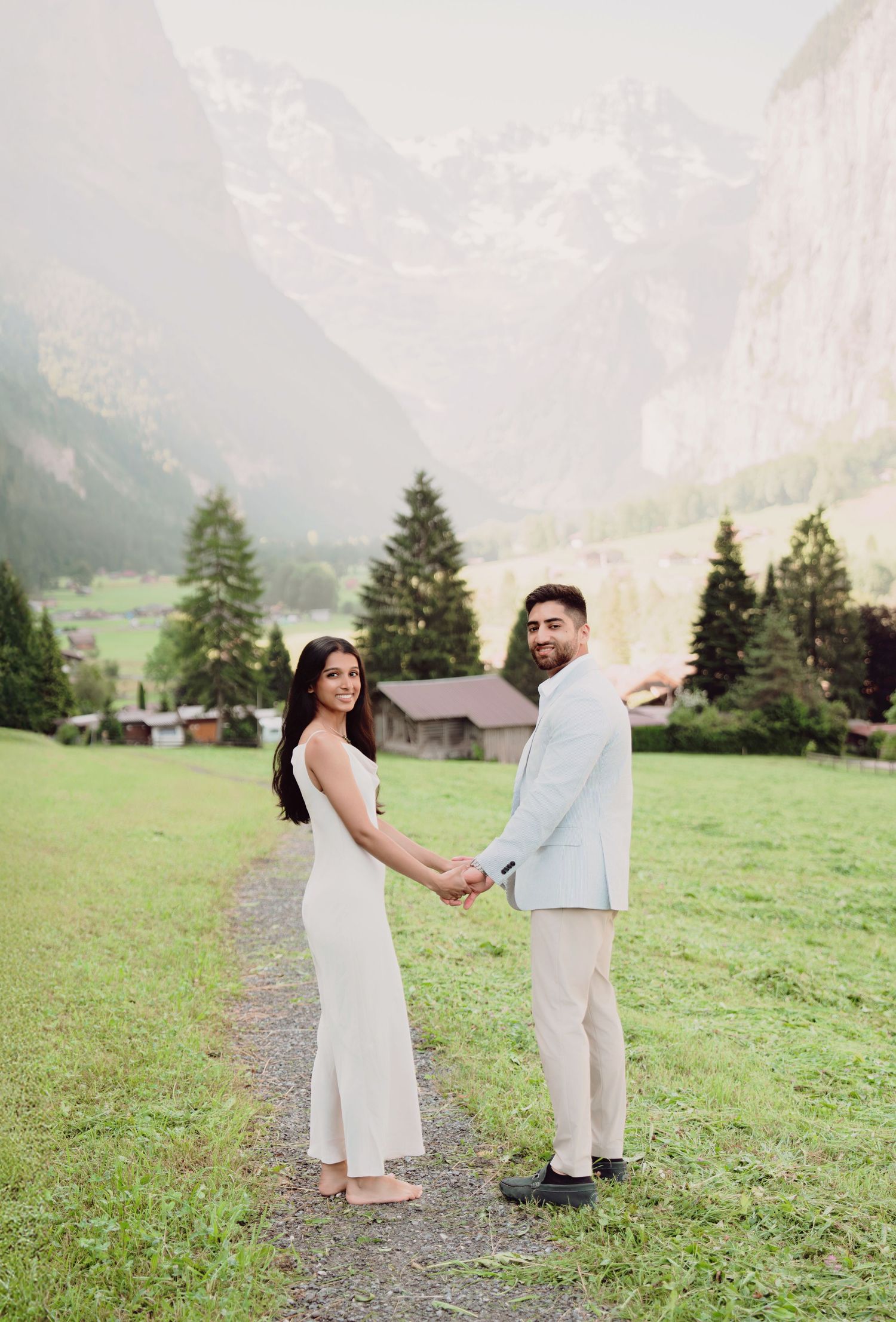 Engaged couple holding hands on a grassy path with stunning Swiss mountain scenery behind them.