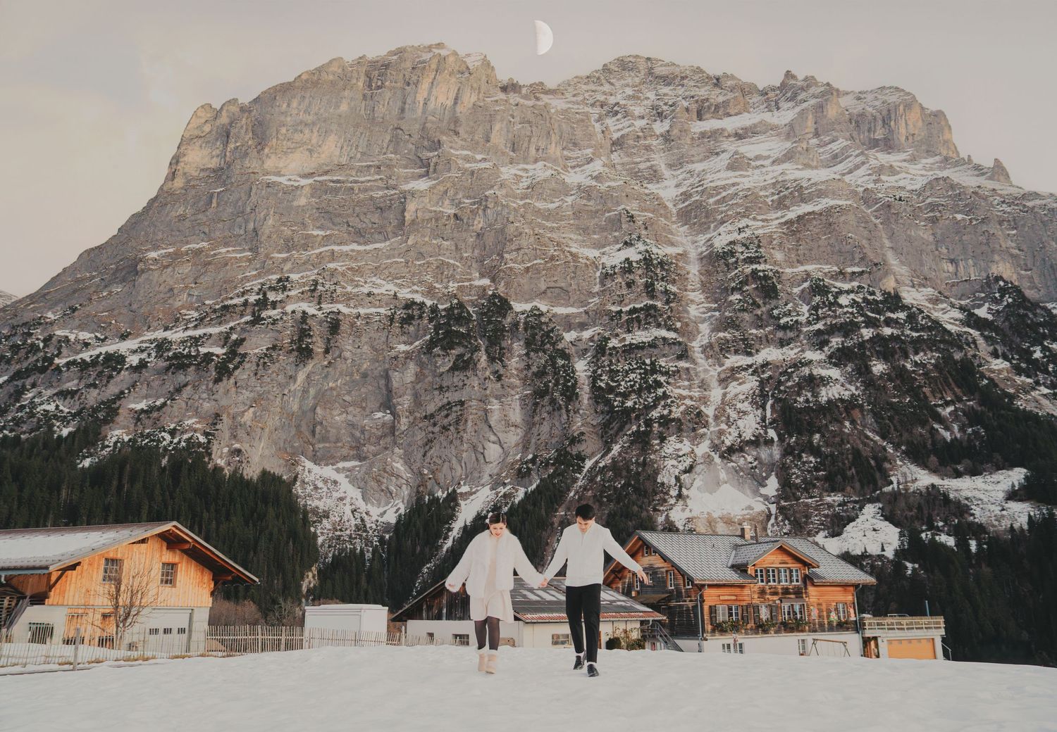 Couple holding hands walking toward snowy Alpine chalets beneath a towering rocky mountain with crescent moon.