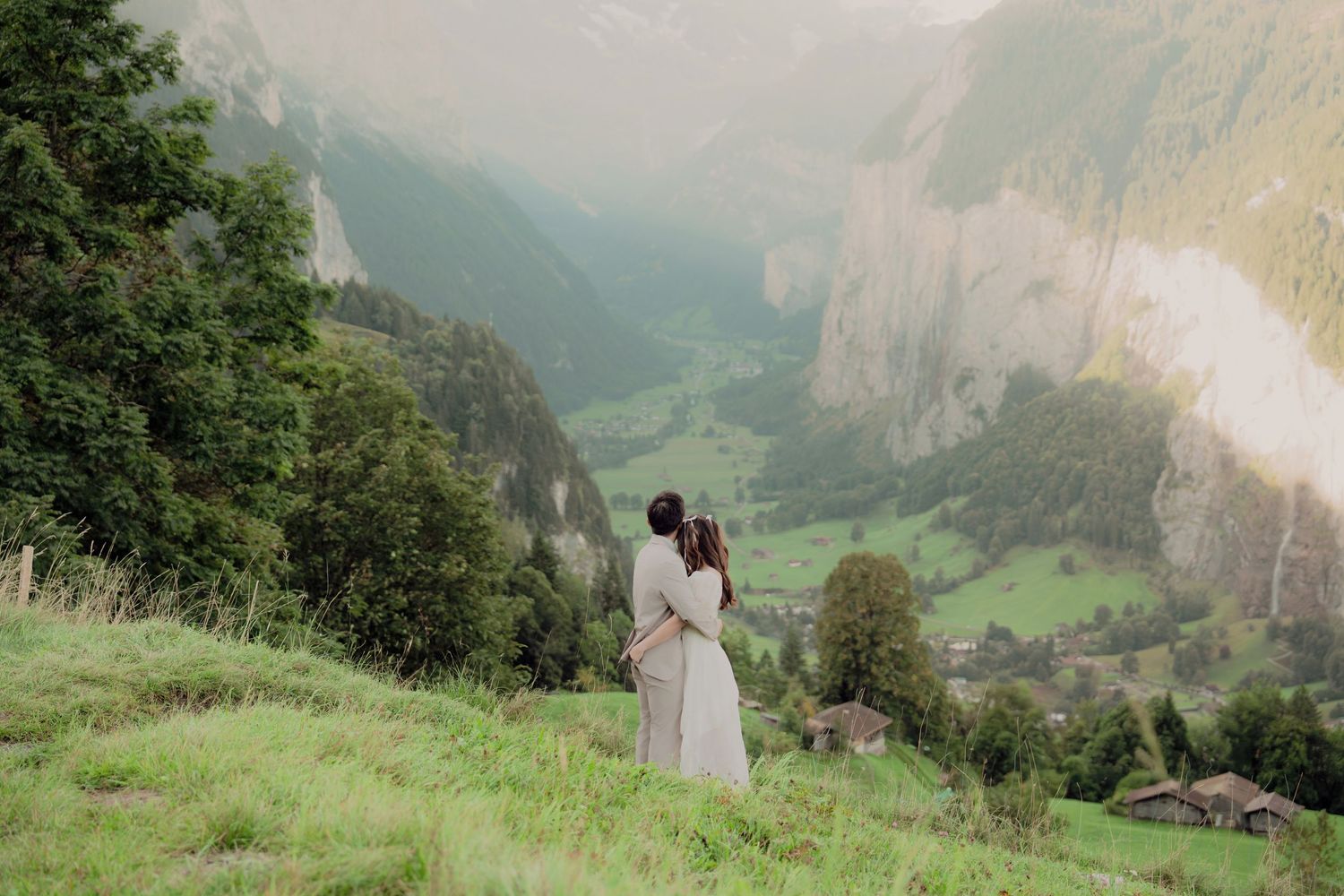 Couple embracing on a grassy Alpine hillside while gazing at a scenic Swiss valley surrounded by towering cliffs.