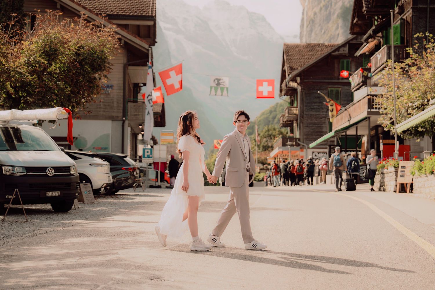 Couple walking hand in hand on sunny Swiss street with national flags, woman in white dress and man in grey suit.