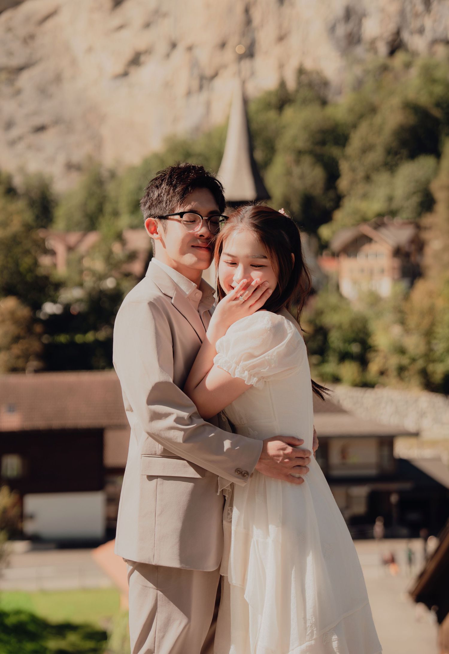 Couple embracing warmly with a charming Swiss village church and rocky cliffs behind them.