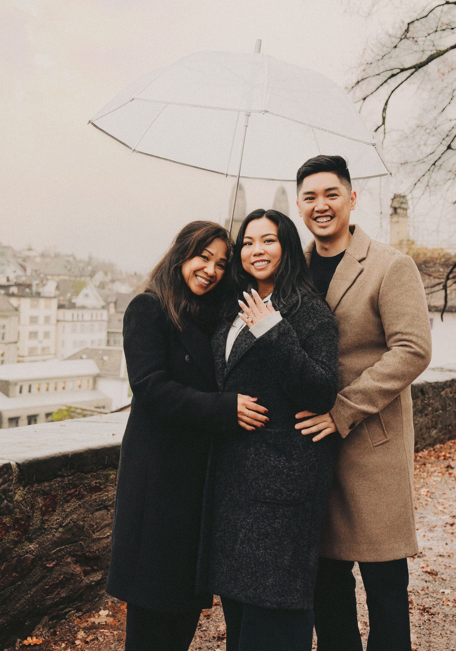 Three friends laugh together under an umbrella, with one showing off an engagement ring in a European city.