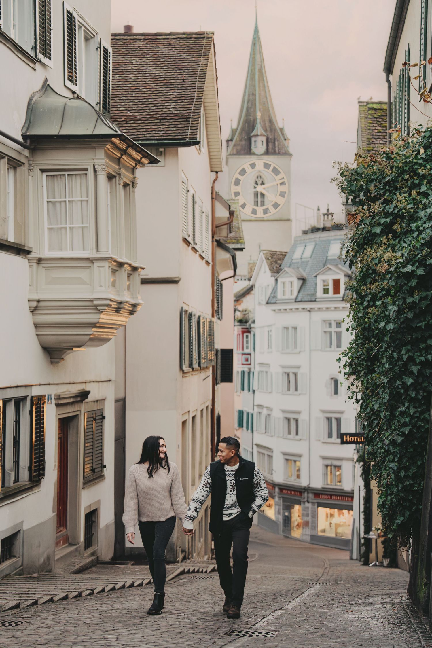 Couple walking hand-in-hand through charming European cobblestone street with clock tower.