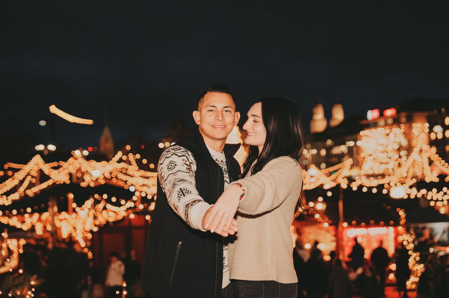 Couple sharing a tender embrace at a warmly lit Christmas market with glowing holiday decorations.