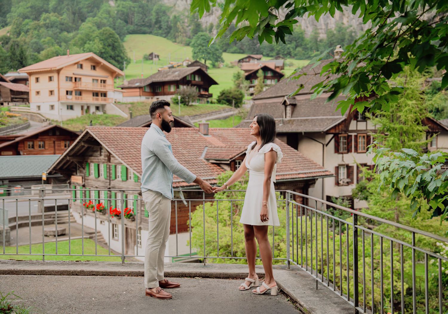 Couple holding hands on a bridge in a charming Swiss Alpine village with traditional chalets on a green hillside.