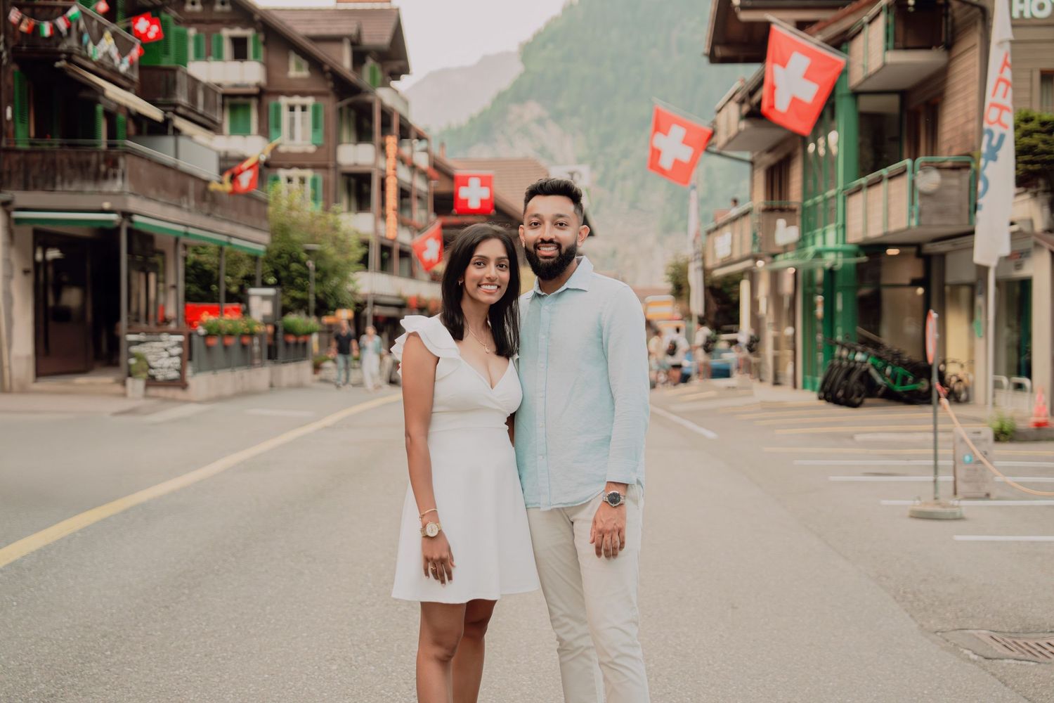 Smiling couple standing together on charming Swiss village main street with traditional buildings and Swiss flags displayed.