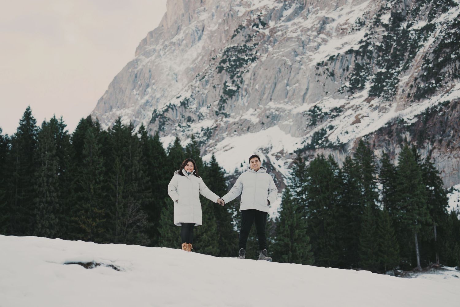 Joyful couple running hand in hand through snow with pine trees and mountain behind.