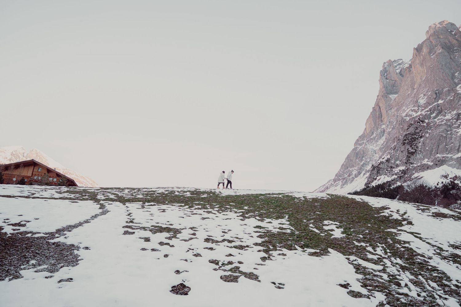 Two people walking across snowy alpine meadow with rocky mountain and rustic hut nearby.