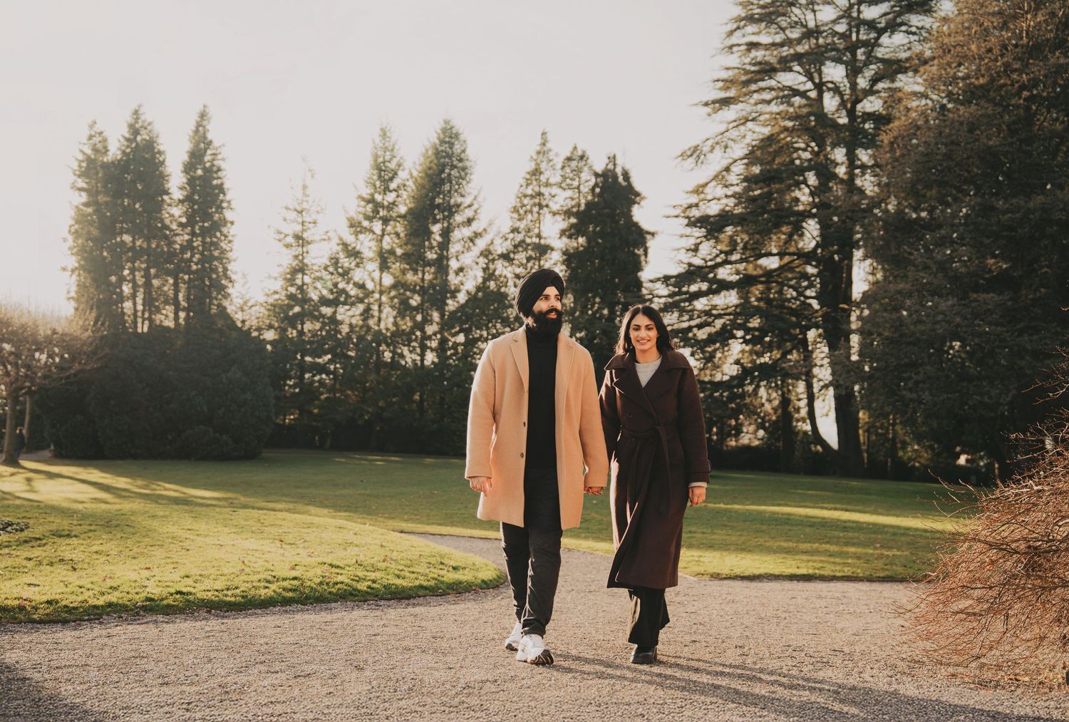 Couple walking hand in hand on a garden path surrounded by tall trees in soft winter light.
